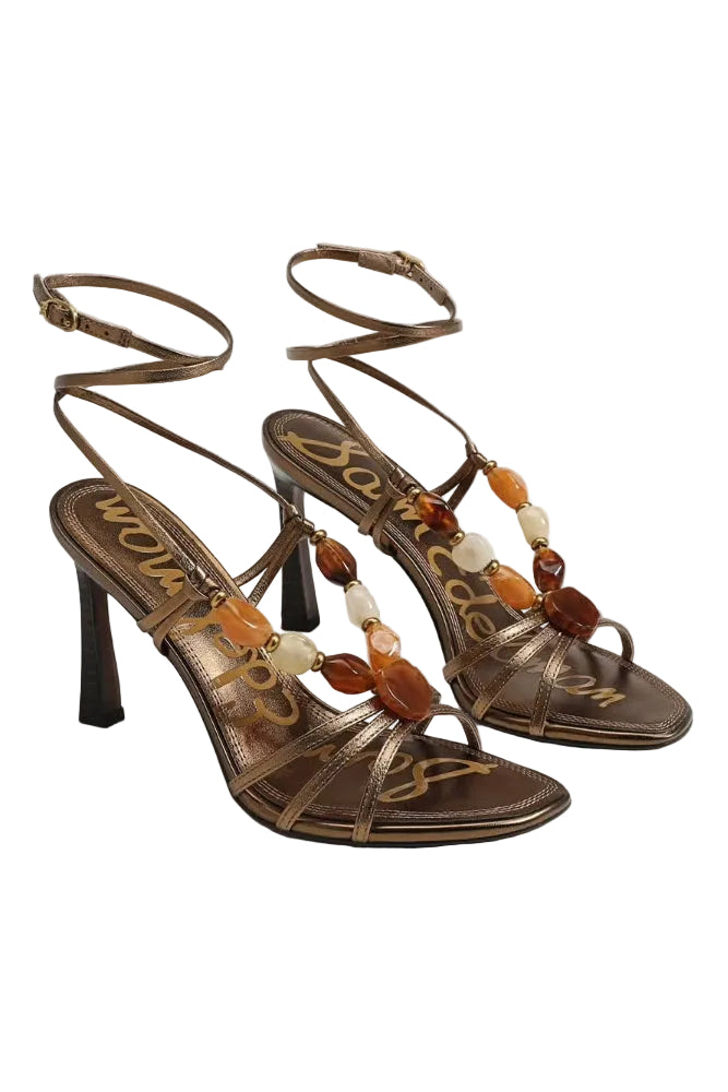 A pair of metallic bronze strappy high-heeled sandals with a beaded ankle strap featuring amber, orange, and cream beads, shown on a light gray background.