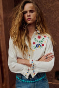 A woman with long, wavy light brown hair stands with her arms crossed, wearing a white long-sleeve button-up shirt decorated with colorful floral embroidery on the chest and cuffs, paired with blue jeans and a turquoise beaded belt. She is posed against a textured, warm brown wall.
