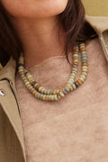 A close-up of a person wearing a double-strand beaded necklace made of natural stone beads in muted earthy tones, styled with a beige knit top and an olive green jacket.