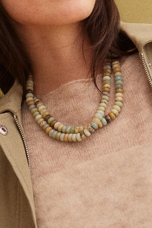 A close-up of a person wearing a double-strand beaded necklace made of natural stone beads in muted earthy tones, styled with a beige knit top and an olive green jacket.