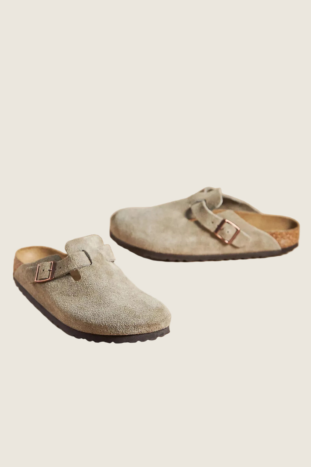 A pair of light taupe suede clogs with double straps and metal buckles, displayed on a solid beige background.