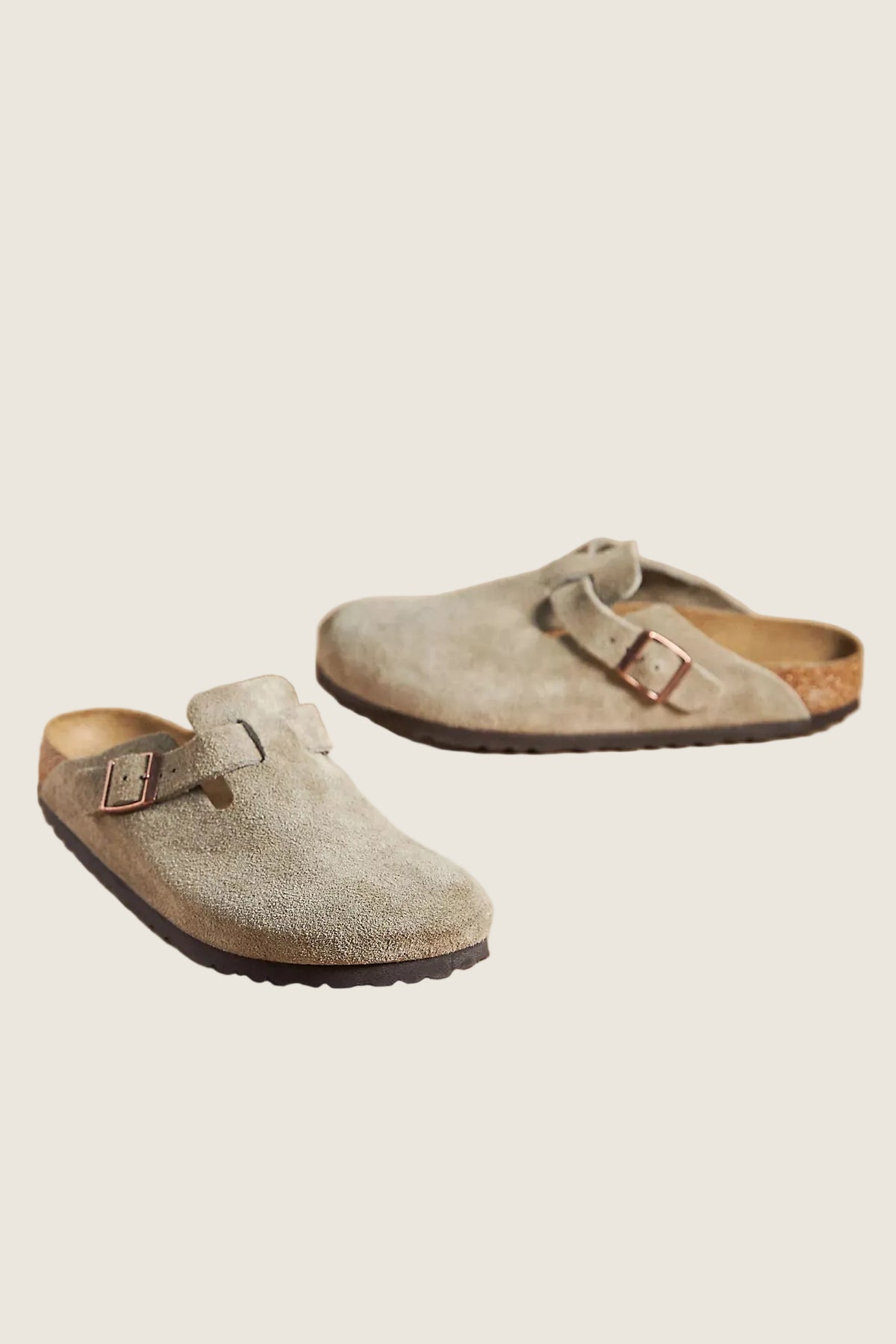 A pair of light taupe suede clogs with double straps and metal buckles, displayed on a solid beige background.