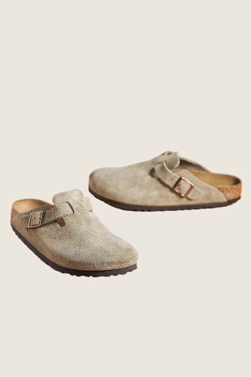 A pair of light taupe suede clogs with double straps and metal buckles, displayed on a solid beige background.