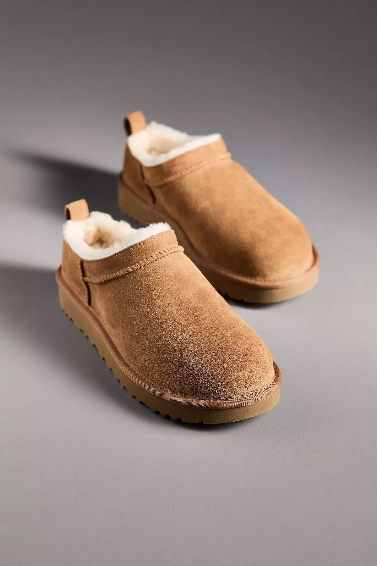 A pair of tan suede slip-on boots with white shearling lining, displayed on a neutral gray background.