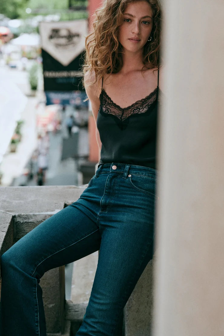A woman wearing a black lace-trim camisole top and dark blue jeans, leaning against a stone wall in an outdoor urban setting.