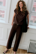 A woman modeling a dark brown short-sleeve top and matching wide-leg pants with decorative side studs, standing in a bright room with white walls and windows.