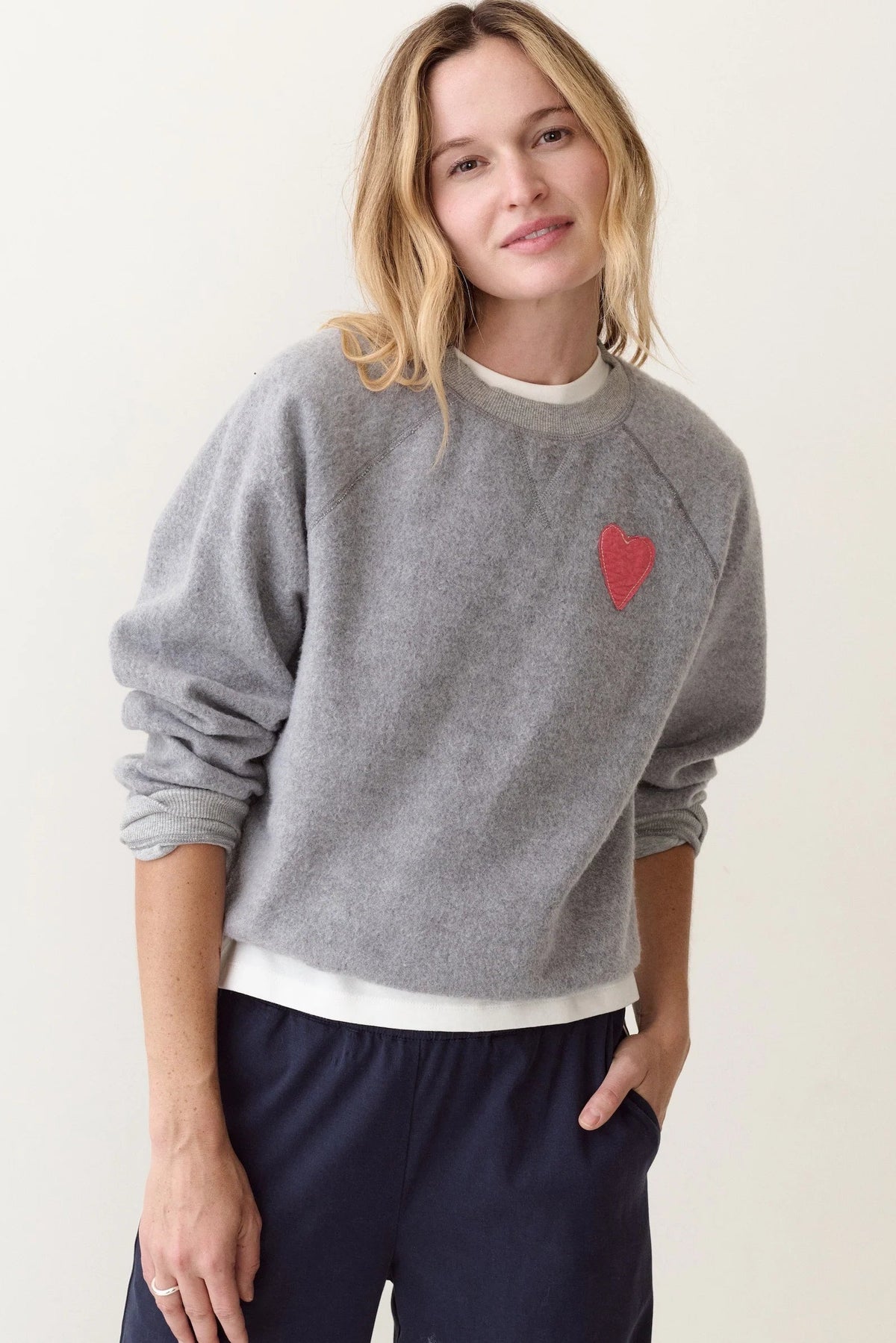 A woman wearing a heather gray crewneck sweatshirt with a small red heart on the chest, layered over a white t-shirt and paired with navy blue sweatpants, set against a plain off-white background.