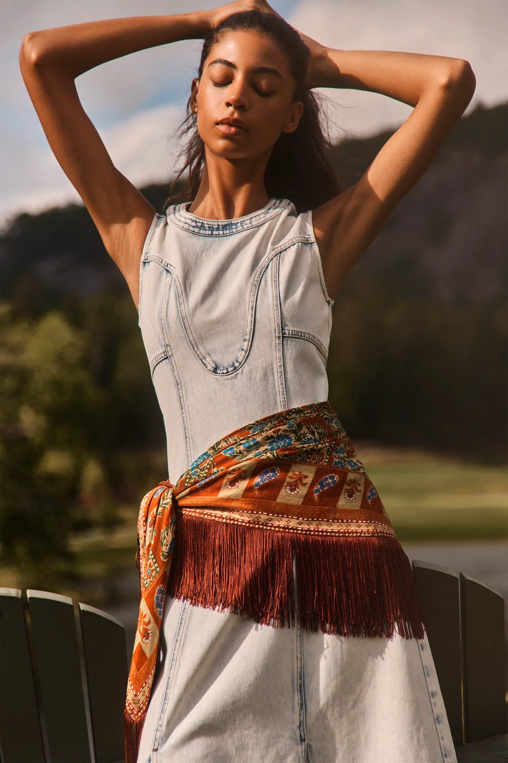A woman outdoors wearing a sleeveless light-wash denim dress with decorative stitching, a rust-colored fringed sash, and a patterned orange scarf tied around her waist. She has her hands behind her head, with blurred trees and hills in the background.