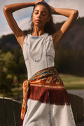 A woman outdoors wearing a sleeveless light-wash denim dress with decorative stitching, a rust-colored fringed sash, and a patterned orange scarf tied around her waist. She has her hands behind her head, with blurred trees and hills in the background.