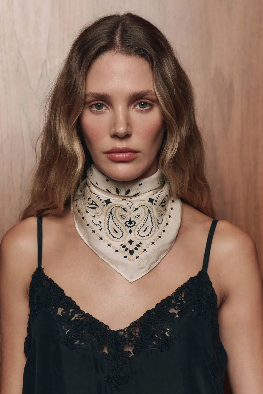 A woman with wavy light brown hair wears a cream-colored paisley bandana tied around her neck over a black lace camisole top, set against a warm wood-paneled background.