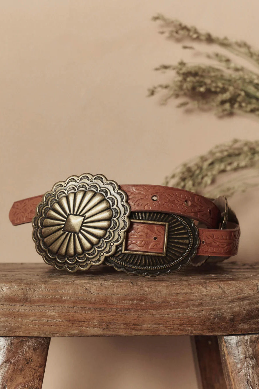 Brown leather belt with a large decorative buckle on a wooden surface.