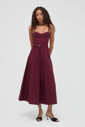 A woman modeling a sleeveless midi dress in deep burgundy with a matching belt and silver buckle, styled with black sandals against a light gray studio background.