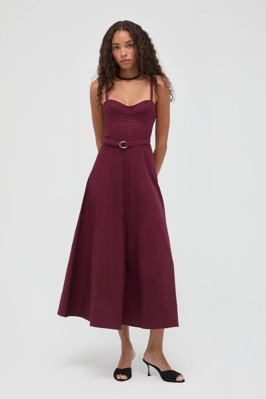 A woman modeling a sleeveless midi dress in deep burgundy with a matching belt and silver buckle, styled with black sandals against a light gray studio background.
