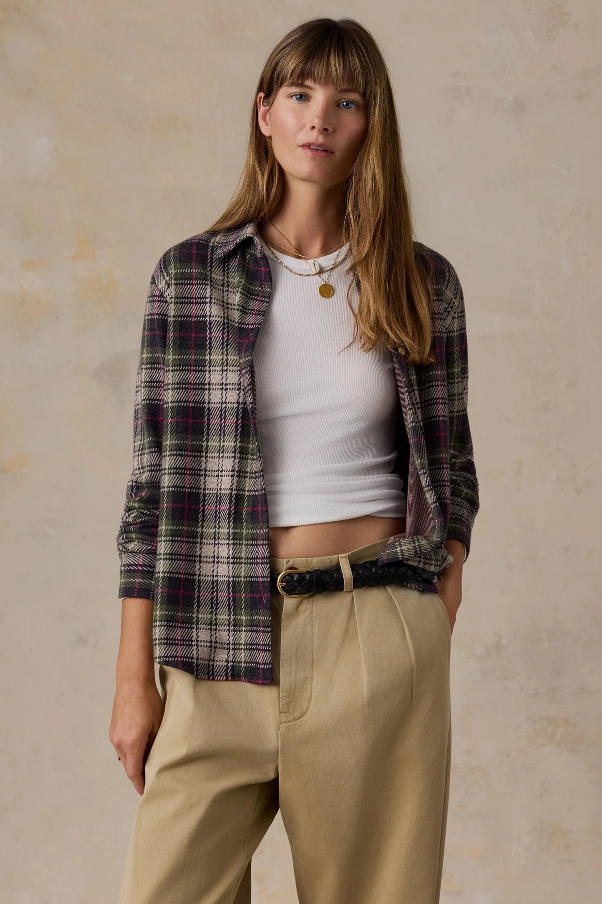 A woman modeling a long-sleeve plaid button-up shirt in dark green, purple, and white, worn open over a white ribbed tank top and paired with beige trousers and a black belt, against a textured beige studio backdrop.