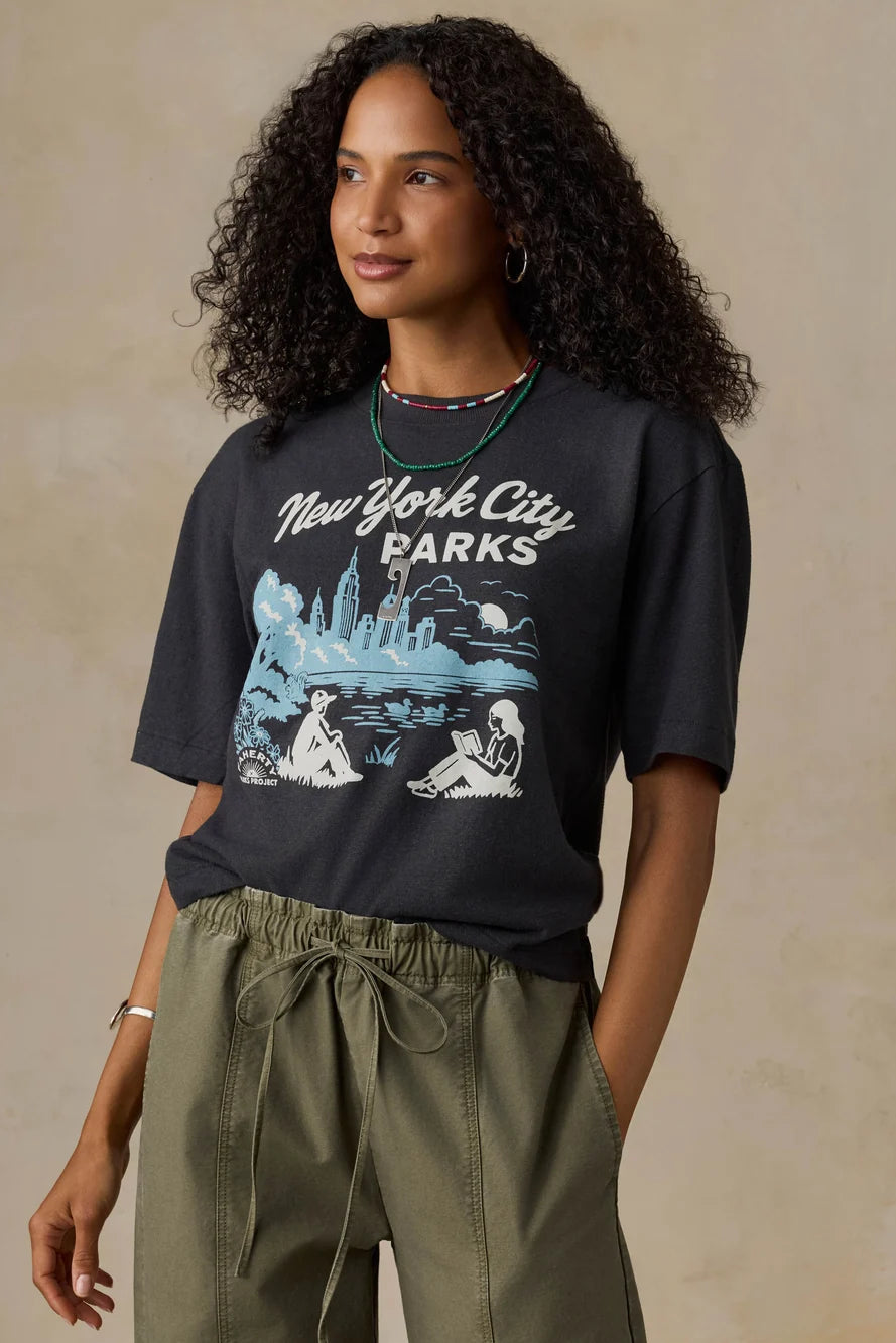 A woman wearing a dark charcoal gray graphic t-shirt that reads 'New York City PARKS' with a blue and white illustration, paired with olive green drawstring pants, set against a textured beige background.