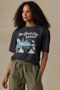 A woman wearing a dark charcoal gray graphic t-shirt that reads 'New York City PARKS' with a blue and white illustration, paired with olive green drawstring pants, set against a textured beige background.
