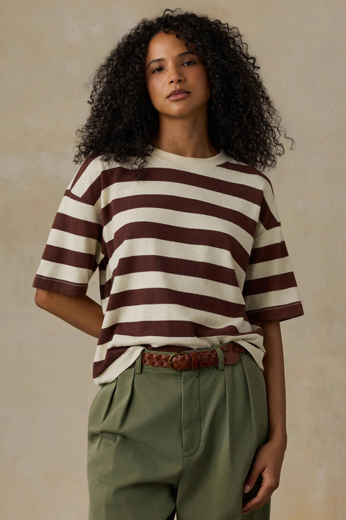 A woman with curly dark hair wearing a brown and cream horizontally striped short-sleeve t-shirt, olive green trousers, and a brown woven belt, standing against a textured beige background.