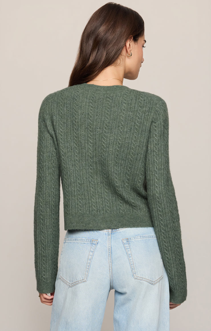 A woman viewed from the back wearing a sage green cable-knit cropped sweater and light-wash blue jeans against a plain beige studio background.