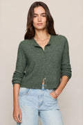 A woman modeling a dark green cable-knit cropped button-up cardigan with light-wash blue jeans, standing against a solid light beige background.