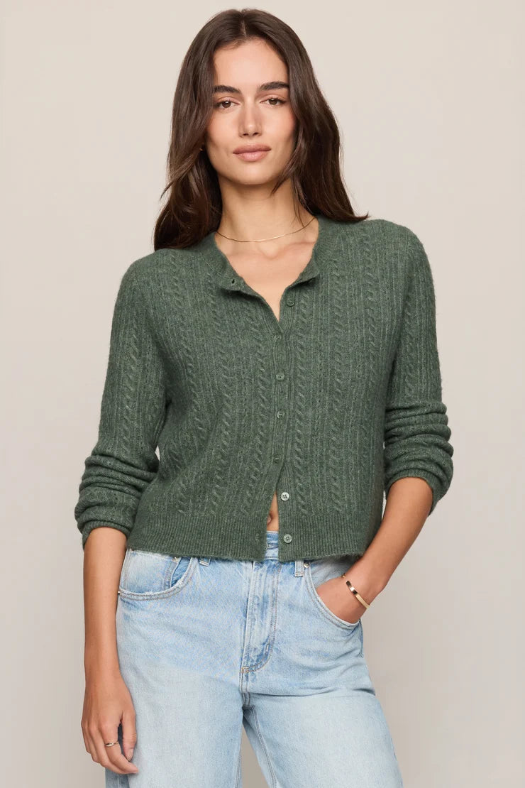 A woman modeling a dark green cable-knit cropped button-up cardigan with light-wash blue jeans, standing against a solid light beige background.