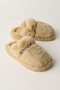 A pair of beige faux fur slippers with a decorative silver rhinestone embellishment across the top, displayed on a clean off-white background.