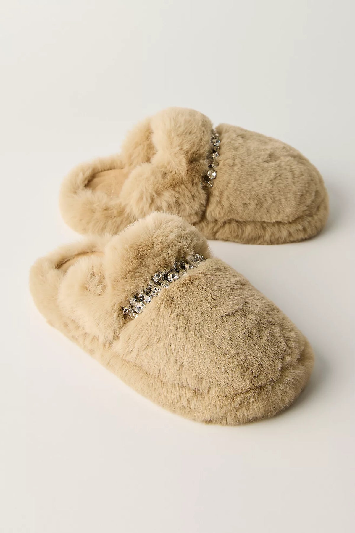 A pair of beige faux fur slippers with a decorative silver rhinestone embellishment across the top, displayed on a clean off-white background.