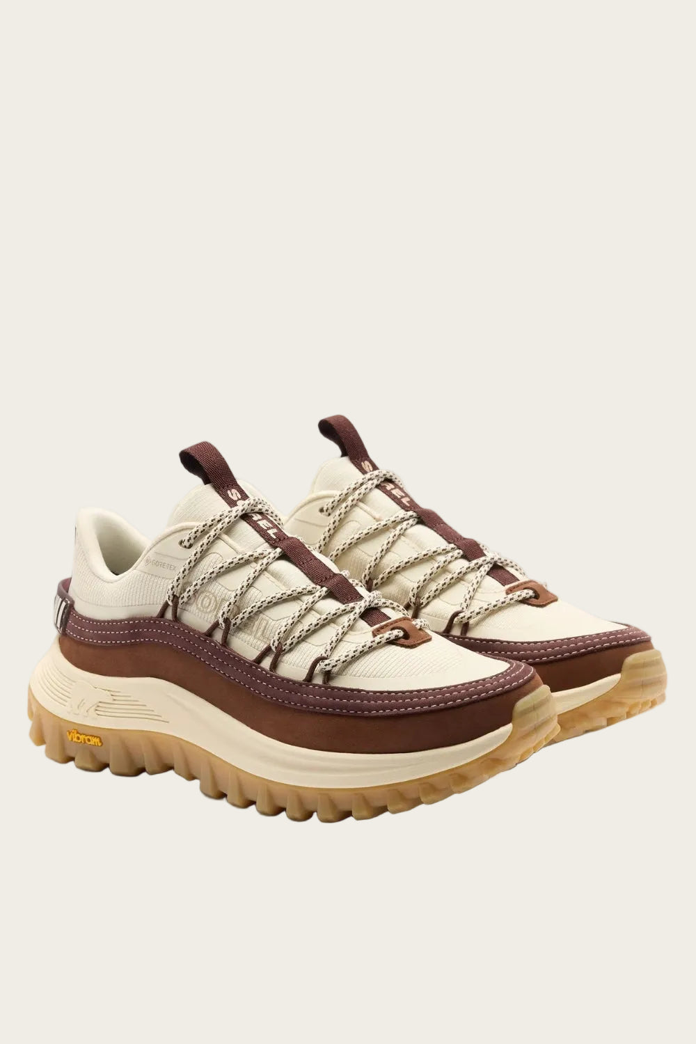 A pair of rugged platform sneakers in cream, brown, and beige, featuring chunky laces, a thick sole, and Vibram branding on the toe and heel.